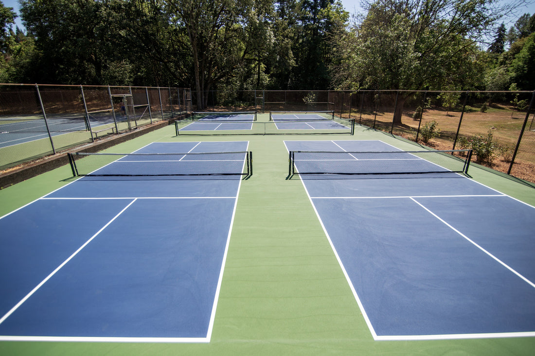 Four outdoor pickleball courts with blue surfaces and white lines, surrounded by green areas and chain-link fences, set against a backdrop of dense trees.